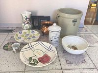 View of lot showing two Fitz and Floyd ceramic plates, large stoneware crock, ceramic vase, copper pot, lidded cafe jar, decorative bowl, and small creamer cup on floor tiles.