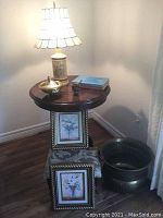 Dark wood round side table holding a ceramic floral lamp with scalloped beige lampshade, small lidded bowl, a tiny wooden item, and two books. Two framed floral prints lean against the table skirt. A large decorative brass bowl is next to the table on the floor.