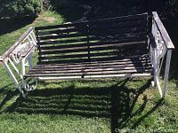 Outdoor metal and wooden garden swing bench showing seating and backrest with horizontal slats and decorative scrollwork sides, photographed in grass.