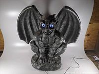 Front view of gray animated gargoyle with blue illuminated eyes and spread wings, placed on white backdrop showing power cord.