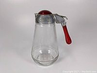 Front and side view of the antique syrup pitcher showing the clear glass body, silver metal lid, and red Bakelite handle and knob.