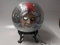 Front view of the spirit ball showing the face inside the clear spherical dome with one eye lit up and blinking, resting on a black claw-foot tripod base.