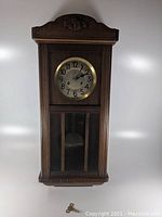 Full front view of wooden German regulator wall clock with pendulum compartment and carved top.