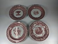 Four ceramic plates with red patterns showing four different designs including an urn, roses, a garden scene, and floral medallions.