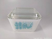 Front angled view of small white Pyrex dish with blue Amish Butterprint rooster and tulips design and clear ribbed glass lid.