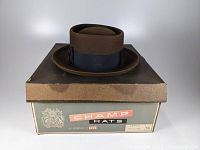 Front view of the chocolate brown felt pork pie hat resting on the vintage Champ Hats cardboard box.