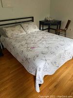 Full view of queen size bed with linens, wooden floor, chair and corner table nearby