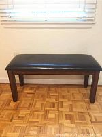 Full view of a padded bench with dark wood legs on a parquet floor under a window with blinds.