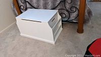 White wooden storage chest with hinged lid and side magazine holders, placed on carpet near bed.