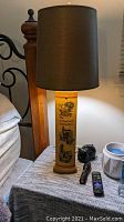 Tall table lamp with a wood base decorated with black drawings of vintage German country scenes and black text, topped with a black lampshade.