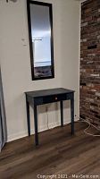 Full view of the black wooden side table under the rectangular mirror on wall in room with dark flooring and brick wall.