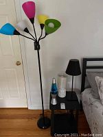 Photo showing the small black side table, floor lamp with multicolored shades, black table lamp, white ceramic base lamp, and silver-blue lava lamp all arranged in bedroom corner.