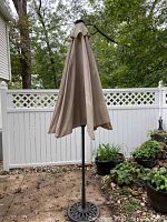 Full view of closed patio umbrella with beige fabric and metal base on outdoor patio surrounded by plants and white fence.