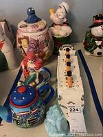 Overview of six ceramic items including Boston teapot, Ariel teapot, Cookie Monster jar, Popeye jar, snowman jar, Titanic jar on table.