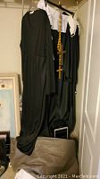Full view of two costumes hanging on hangers, one black robe with a white headpiece and collar (nun costume), the other a black priest robe. Brown fabric or cloth is in the foreground.