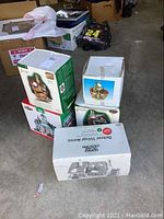 Group photo of boxed Dept 56 lighted buildings stacked on the floor with a cardboard box and miscellaneous items around them.