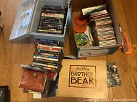 Photo showing a gray container filled with DVDs, a box with CDs and DVDs organized, a small stack of DVDs outside the container, leather wallet, and a Brother Bear Disney poster.