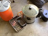 Photo showing large orange cooler, stainless steel electric fryer with glass lid, small metal grilling rack with drip pan.