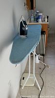 Rowenta iron placed on top of blue fabric-covered ironing board with white metal frame in tiled room.