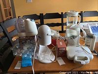 Lot of various kitchen appliances arranged on a wooden table including food processor, blender, hand mixer, teapot, waffle maker, coffee grinder, sno cone maker, and a boxed Twinkies bake set.