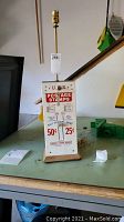 Front view of the vintage postage stamp machine lamp showing pricing and usage details on front panel, lamp socket at top, and wooden base.