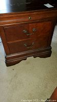 Photo of one of the wooden nightstands showing front view with two drawers and metal handles, some wear and damage on the top surface visible.