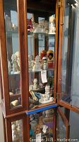 Wooden glass display cabinet containing multiple porcelain figurines and collectibles including Precious Moments characters and other figurines.