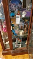Overall view of wooden display case with multiple porcelain figurines and decorations including Precious Moments characters, teddy bear, and mouse figurine.