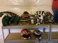 Shelf display showing an assortment of remote controlled toys including a green dinosaur, caveman figure, white and black robots, grey robotic dog, and two plastic helmets below the toys.