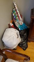 Photo showing multiple rolls of Christmas-themed wrapping paper leaning in corner along with a large roll of white batting and bags containing yarn and other craft materials.