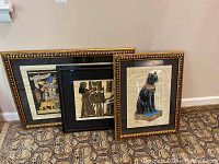Three framed Egyptian papyrus paintings leaning against a wall on carpet, varying in size with detailed golden-black frames.