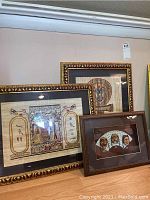 Three framed artworks displayed together: two Egyptian papyrus paintings with gold and black ornate frames and one Asian mask display in a wooden frame.