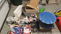Wide shot showing multiple garden decor items including bird feeder, gnomes, cats, garden flags, planters, and galvanized bin on garage floor
