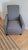 Front view of grey fabric recliner chair with rectangular backrest, armrests, and dark wooden legs on wood floor.