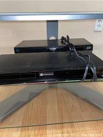 Two Blu-ray players on a glass shelf, Panasonic player visible on top with power cable coiled on it, Sony player underneath.