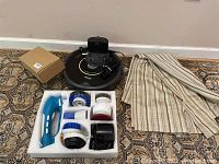 Full lot photo showing Roomba vacuum with charger, handheld brush cleaner with brush attachments in foam holder, black floor buffer, and folded striped drapery.