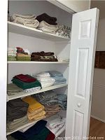 Shelves containing neatly folded bath towels, sheets, tablecloths and napkins in assorted colors including dark grey, blue, beige, mustard yellow and patterned fabrics.