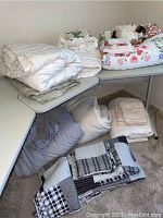 Various comforter sets, quilts, blankets, and linens displayed on tables and floor, including white, floral, and gray items.