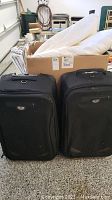 Two black Eagle Creek rolling suitcases standing upright on speckled floor with a cardboard box behind them containing pillows and plastic hangers. Visible scuffs and scratches on suitcases.