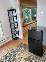 Mini fridge standing on carpeted floor near a doorway, viewed at an angle showing side and front; visible wear and smudges on fridge surface; tall shelving unit with clear plastic barware glasses and bowls in background.