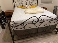 Photo showing black metal bed frame with decorative scrollwork and bedding, with wooden nightstands on either side.
