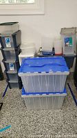 Various stacked plastic storage bins including clear KIS Bi Box with dark lids, larger Iris USA KeepBox storage bins with blue lids (one with duct tape), and smaller clear bins with assorted lids including green and gray. Also includes a red-lidded container possibly for dog food.
