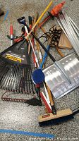 Various long-handled garden tools including rakes, shovels, brooms, squeegee, and metal poles arranged on garage floor.