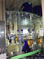Glass bathroom decor items arranged on two shelves inside a mirrored cabinet, showing multiple glass bottles with blue, white, frosted, amber colors and unique stoppers