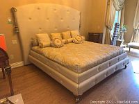 Full view of the king size upholstered bed with headboard, decorative pillows, and patterned bedcover in cream and gold tones in a bedroom setting.