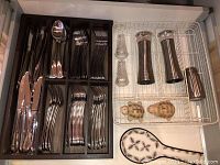 Photo showing wooden cutlery tray with knives, forks, and spoons neatly arranged in compartments alongside metal tray with salt and pepper grinders, glass and ceramic shakers, and ceramic spoon rest.