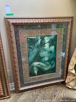 Front view of the wooden ornate frame including glass, matting, and artwork within, resting against a wall on a wooden floor.