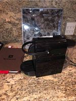 Photo of black Nespresso coffee maker on countertop with pastry cake plate and server box visible behind it, including Nespresso manual booklet