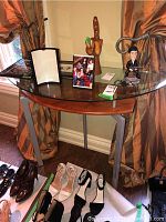 Demi lune table shown with various decorative items on top and footwear displayed underneath to illustrate size and style. Glass top is semi-circular with wooden trim and three metal legs.