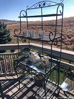 Full view of black metal 4-tier bakers rack on outdoor deck with scenic background. Shows overall structure and shelves with sample decorative items not included.